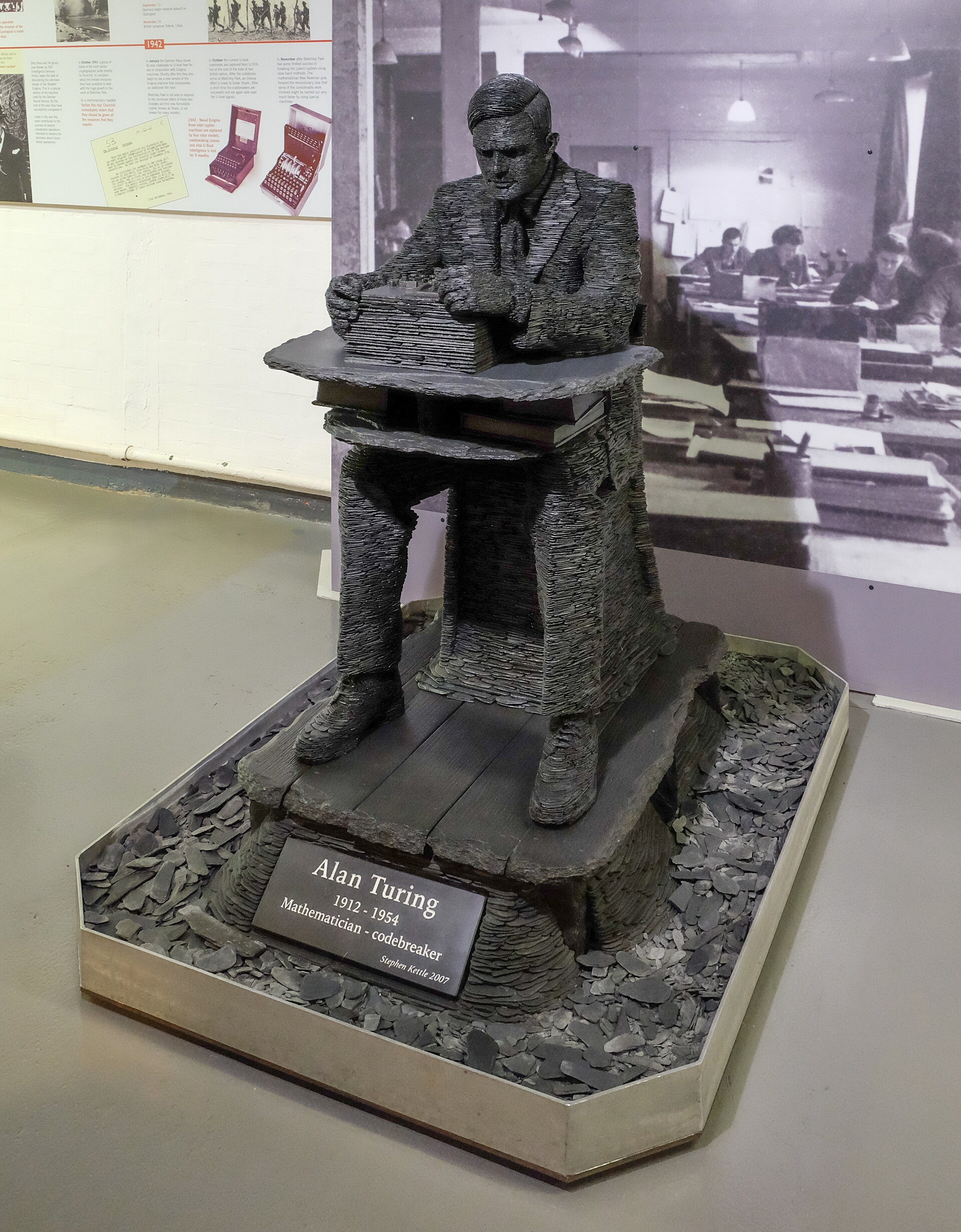 Slate sculpture of Alan Turing seated at a desk, displayed at Bletchley Park with a plinth reading Alan Turing 1912-1954, mathematician and codebreaker